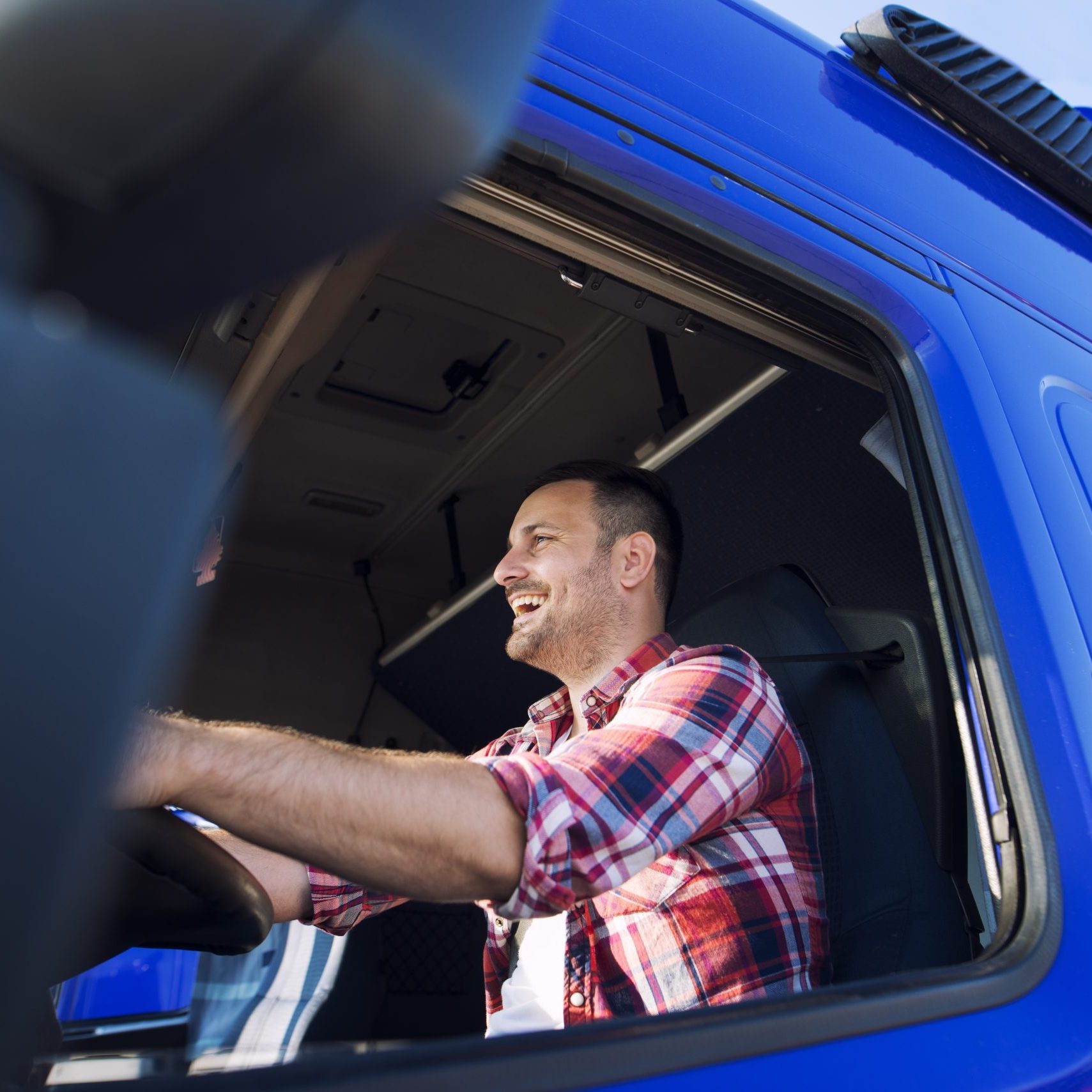 Professional middle aged trucker in cabin driving truck and smiling.